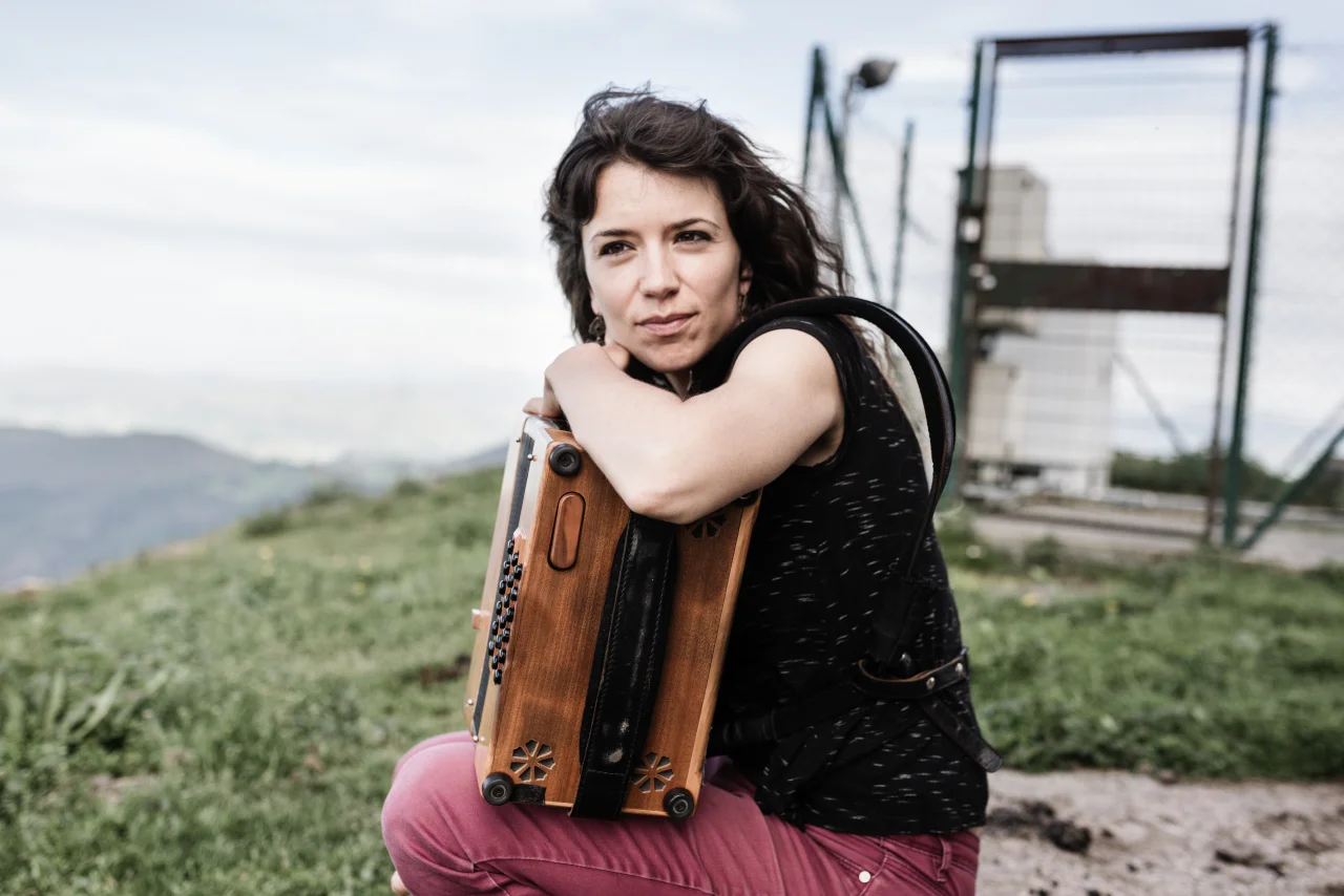 A woman with dark wavy hair sits atop a mountain. She is in profile, but her face is turned to the camera. She is wearing a black sleeveless top and dark pink jeans, and her left arm is hooked over the top of her melodeon, which is resting on her knees. There is grass and a metal fence int he background.