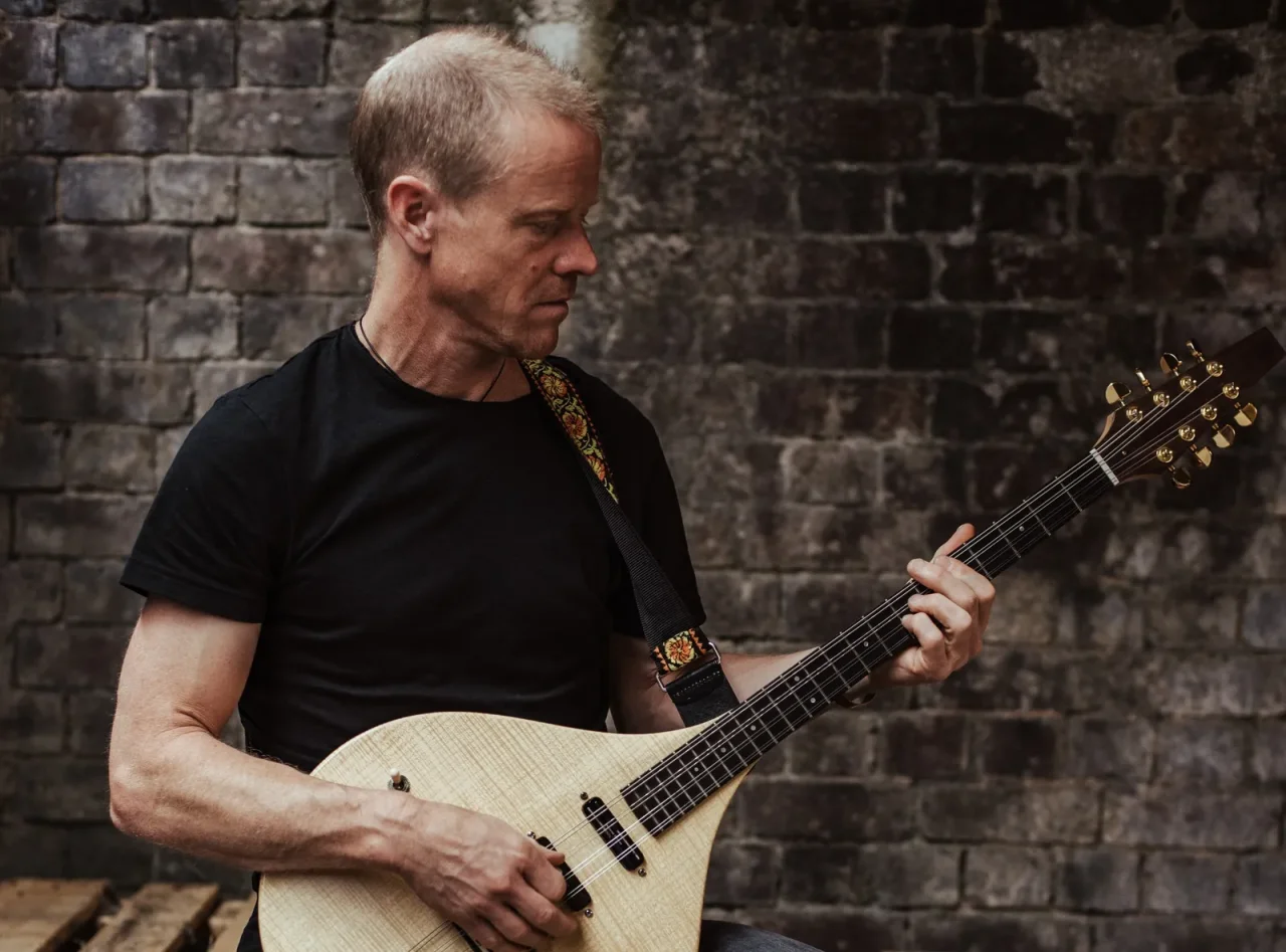 Benji is wearing a black t-shirt and holding his bouzouki. He is looking down at his hand on the frets of the instrument. The bouzouki strap has red and yellow flowers embroidered on it. He is standing in front of a dark brick wall.
