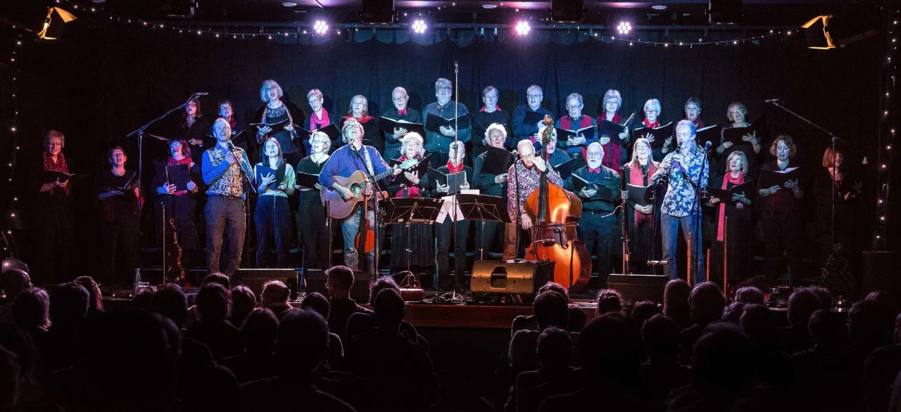The Carnival Band on stage , lit in blue, backed by a 30-strong choir in front of an audience in darkness.