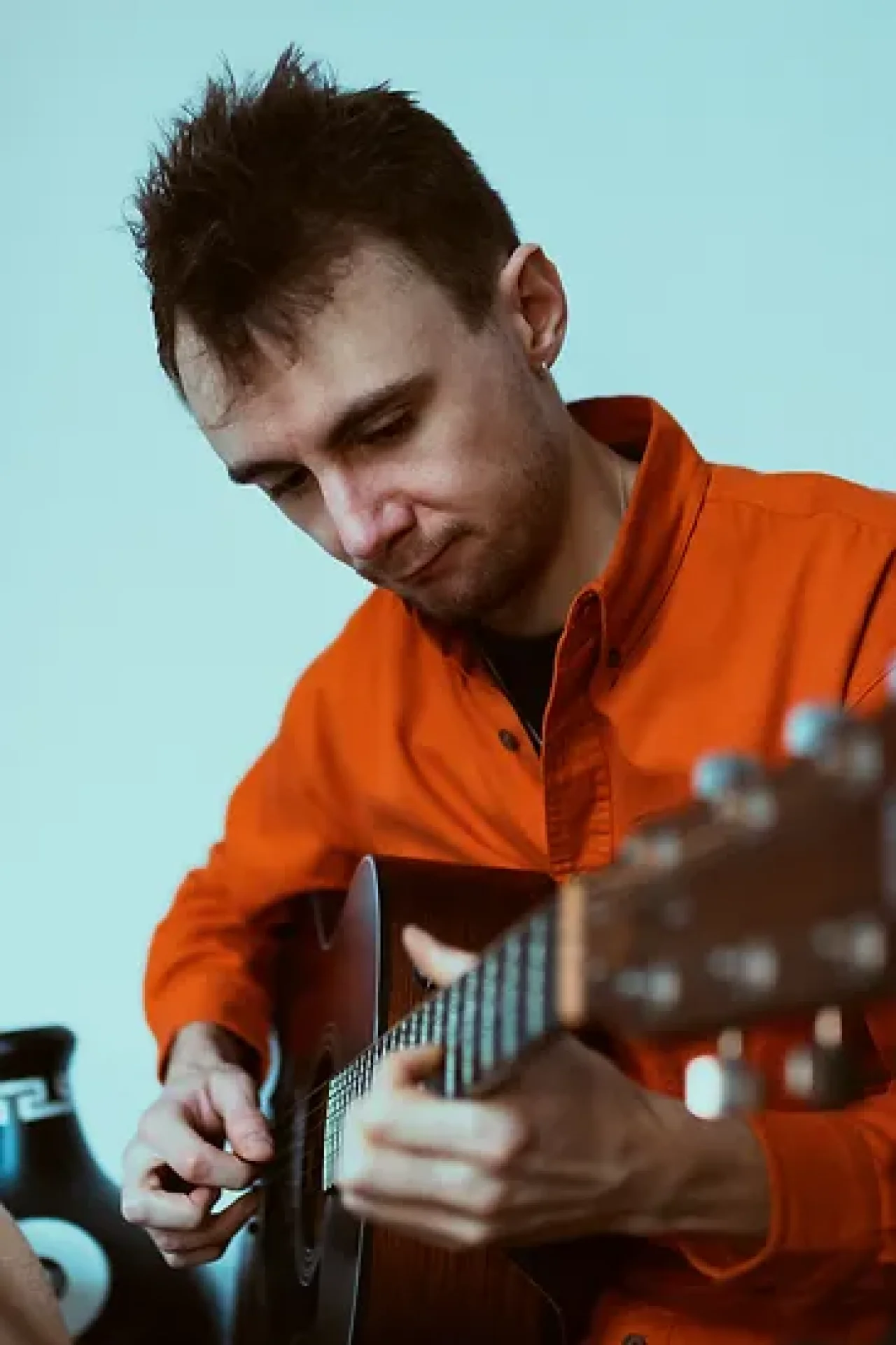Ford plays the guitar. He wears a bright orange shirt with a pale blue background.