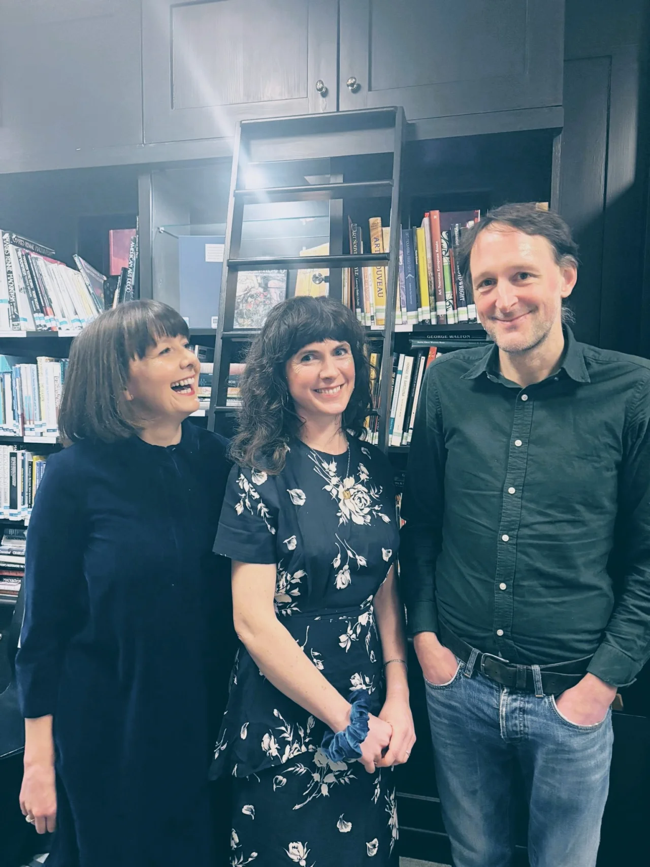 Three members of the Furrow Collective standing in front of a wall of books. They smile and laugh and pose casually.