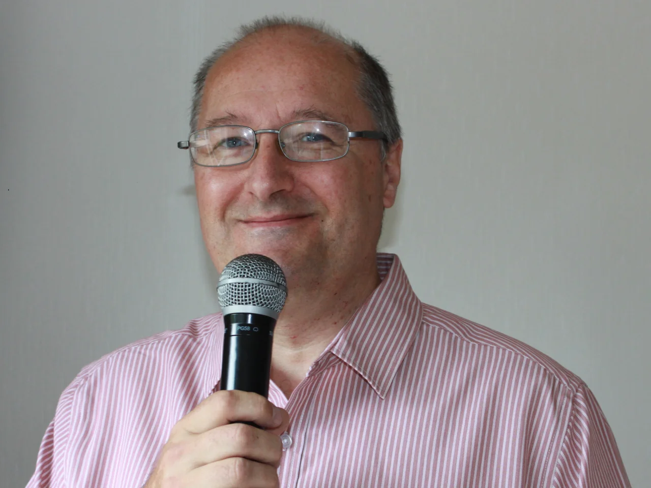 Geoff is wearing a pink and white striped shirt, and holding a microphone up to his mouth. He has metal rimmed glasses and is smiling at the camera.