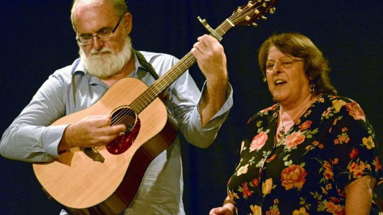 Bob & Gill Berry, Bob plays the guitar whilst Gill sings alongside. Bob wears a blue collared shirt and Gill wears a floral blouse. Both are wearing spectacles.
