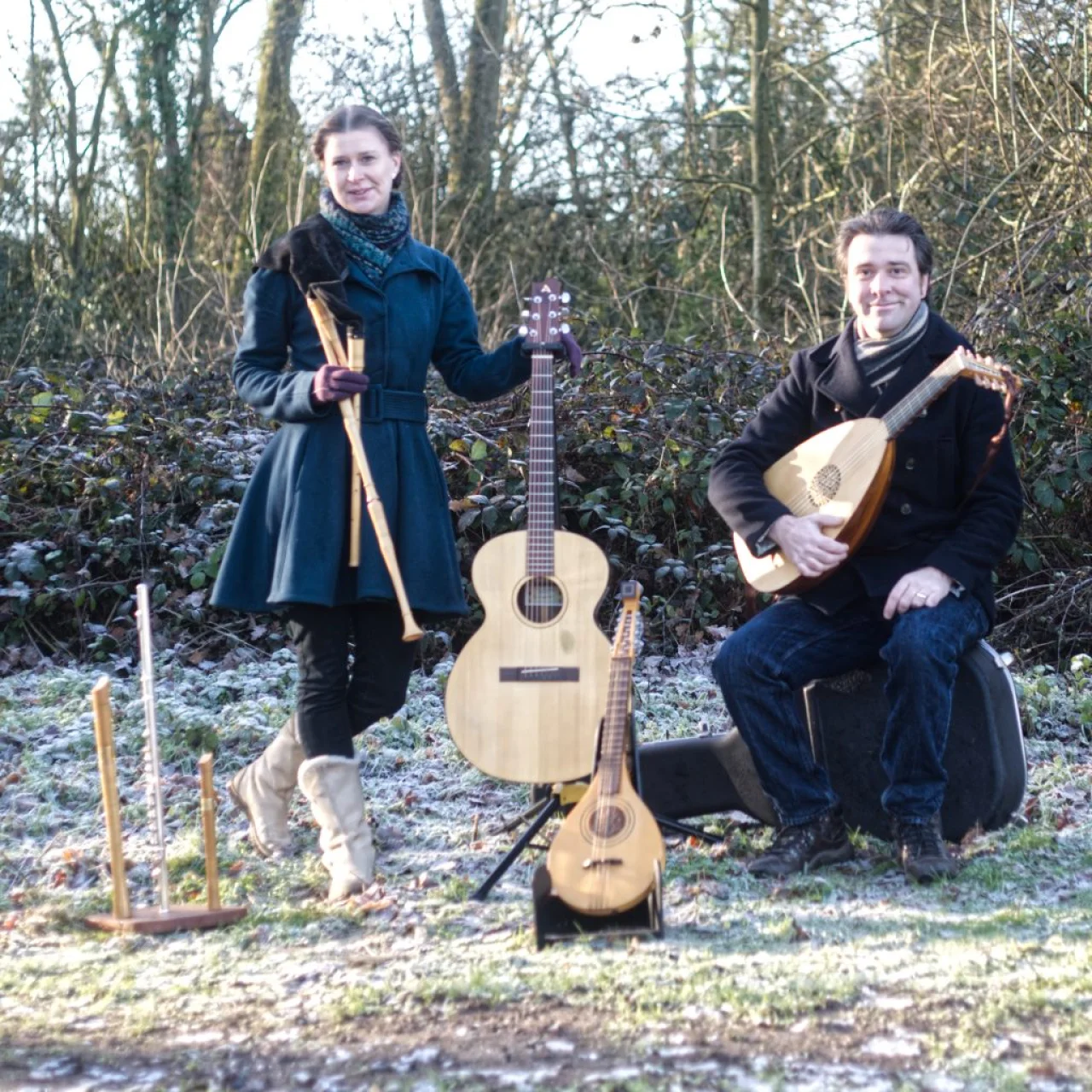 The members of GreenMatthews posed with instruments outside on grass with a hedge and trees in the background. The weather in sunny but cold, the ground has a sprinkle of snow and Sophie and Chris wear warm coats.