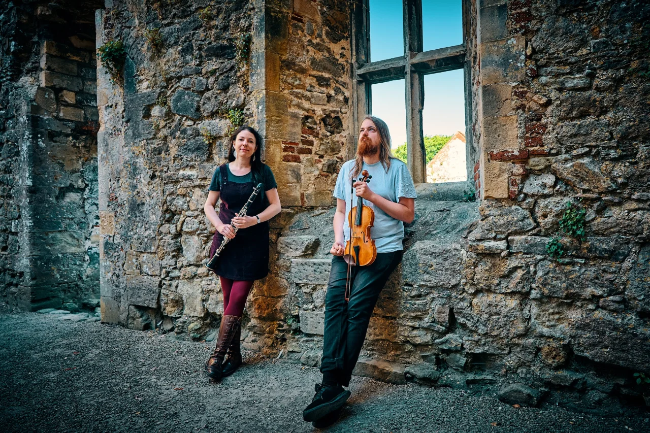 Helen and Lewis are standing in an old building, in front of a stone window with no glass. Helen is wearing a pinafore dress with cool pink tights and brown lace up boots, and is holding a clarinet. She has long dark hair and big earrings and is looking at the camera. Lewis is leaning against the windowsill, holing his violin and bow against his leg. He is wearing a light blue t shirt and dark jeans, and he has long hair and an impressive beard.