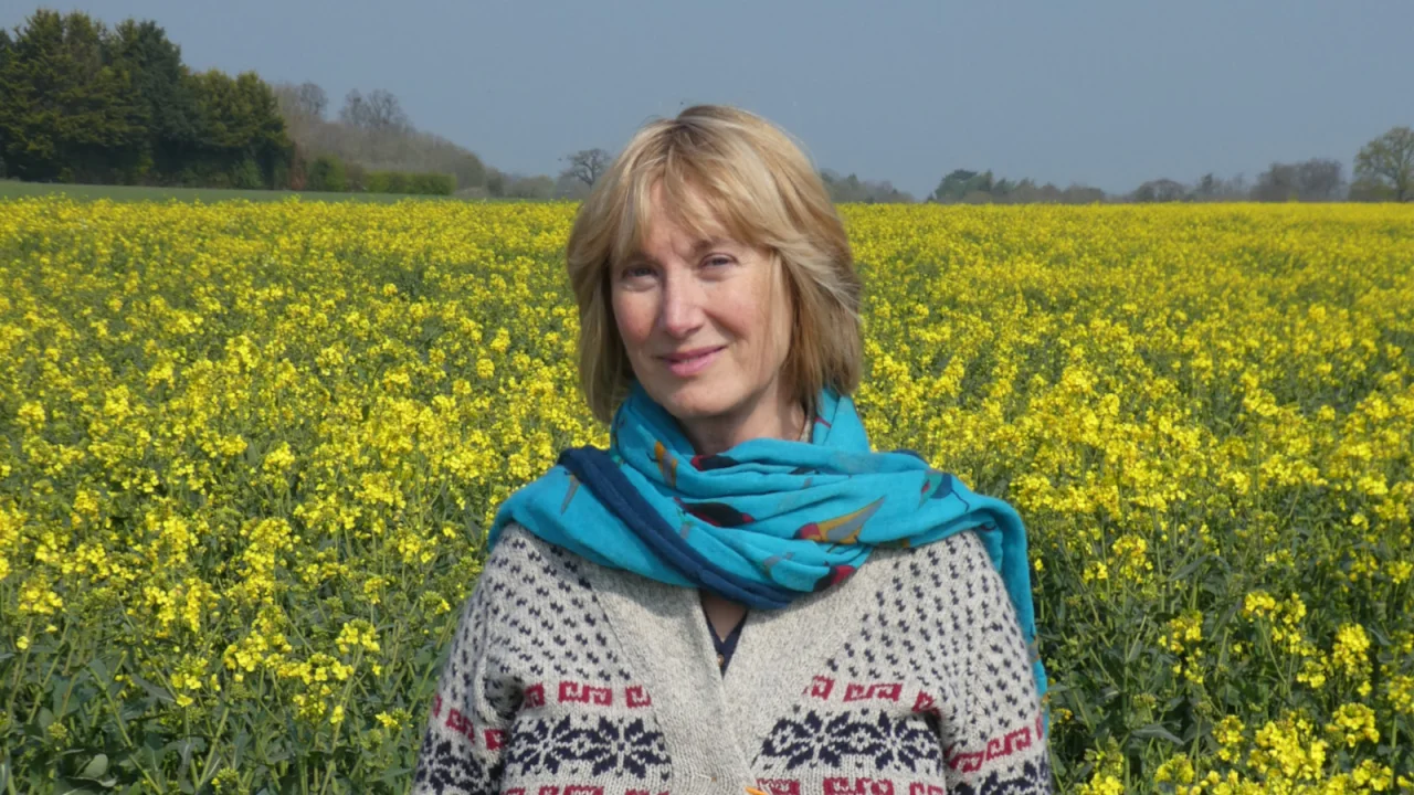 Hilary stands centrally in front of a field of yellow flowers. The sky is blue. Hilary is smiling as she looks into the camera.