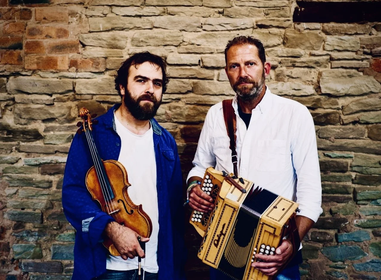 James and Saul hold their instruments whilst looking into the camera. They are photographed from the waist up and are standing in front of a stone wall.