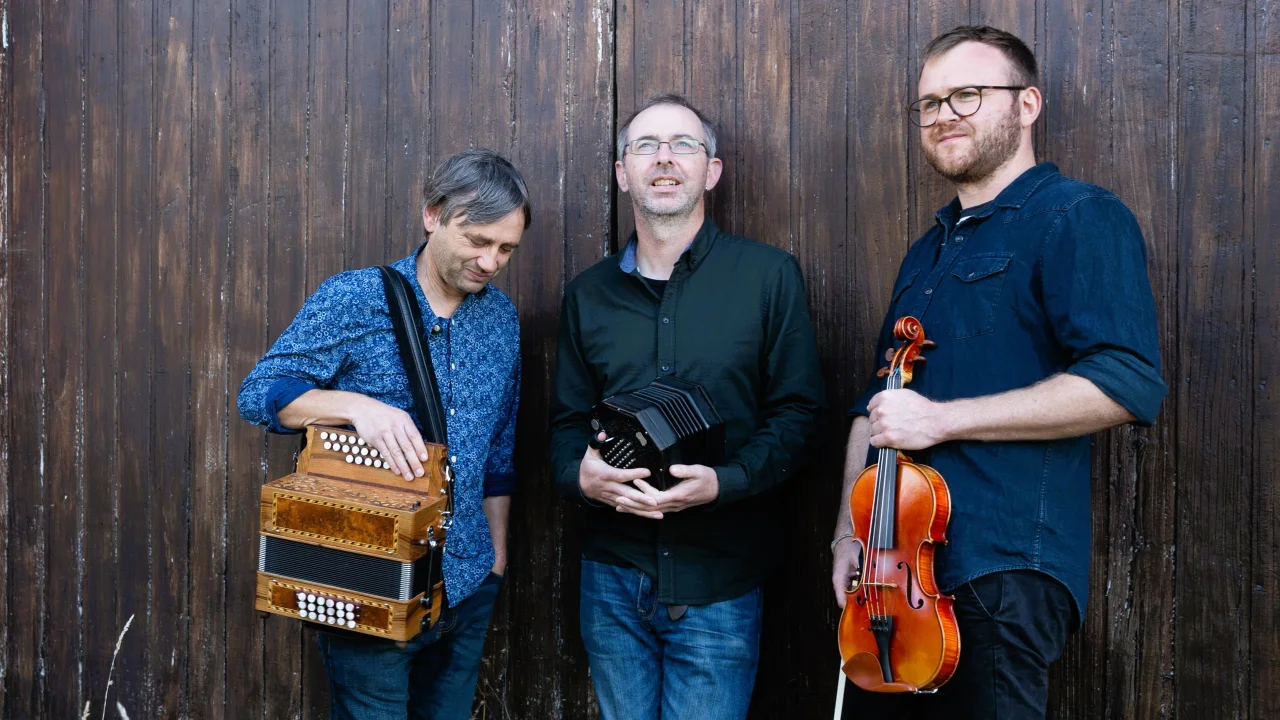 Three men stand against a dark brown wood background. They are holding instruments and none are looking at the camera. On the left, a man with floppy dark grey hair wearing a blue patterned shirt holds a melodeon on a strap over one shoulder and looks at the ground. In the middle, a man in thin-framed glasses and wearing a black shirt and blue jeans holds a black concertina with silver buttons and looks over the photographer's head. On the right, a man in thick-rimmed glasses with a short beard, wearing a dark blue denim shirt and holding a violin, looks over to the left.