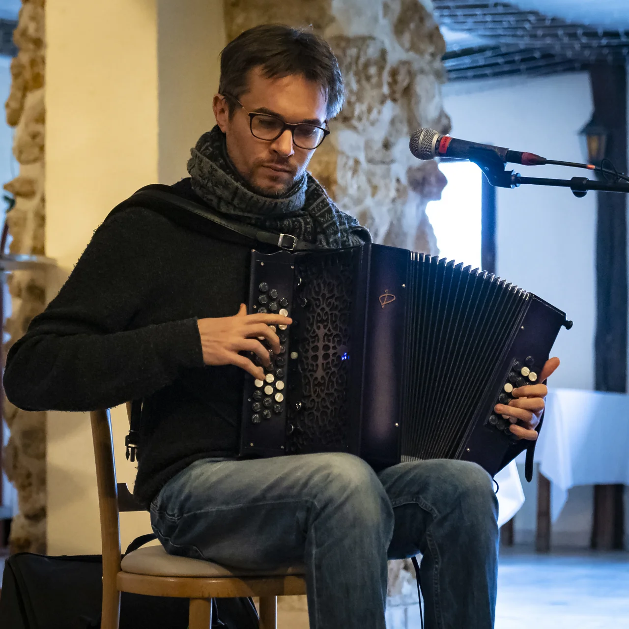 A man with glasses and a short beard sits on a wooden chair and is playing the melodeon. It is a dark purple instrument with black and white buttons. There is a microphone on the right hand side of the photo and the room behind has rugged stone walls.