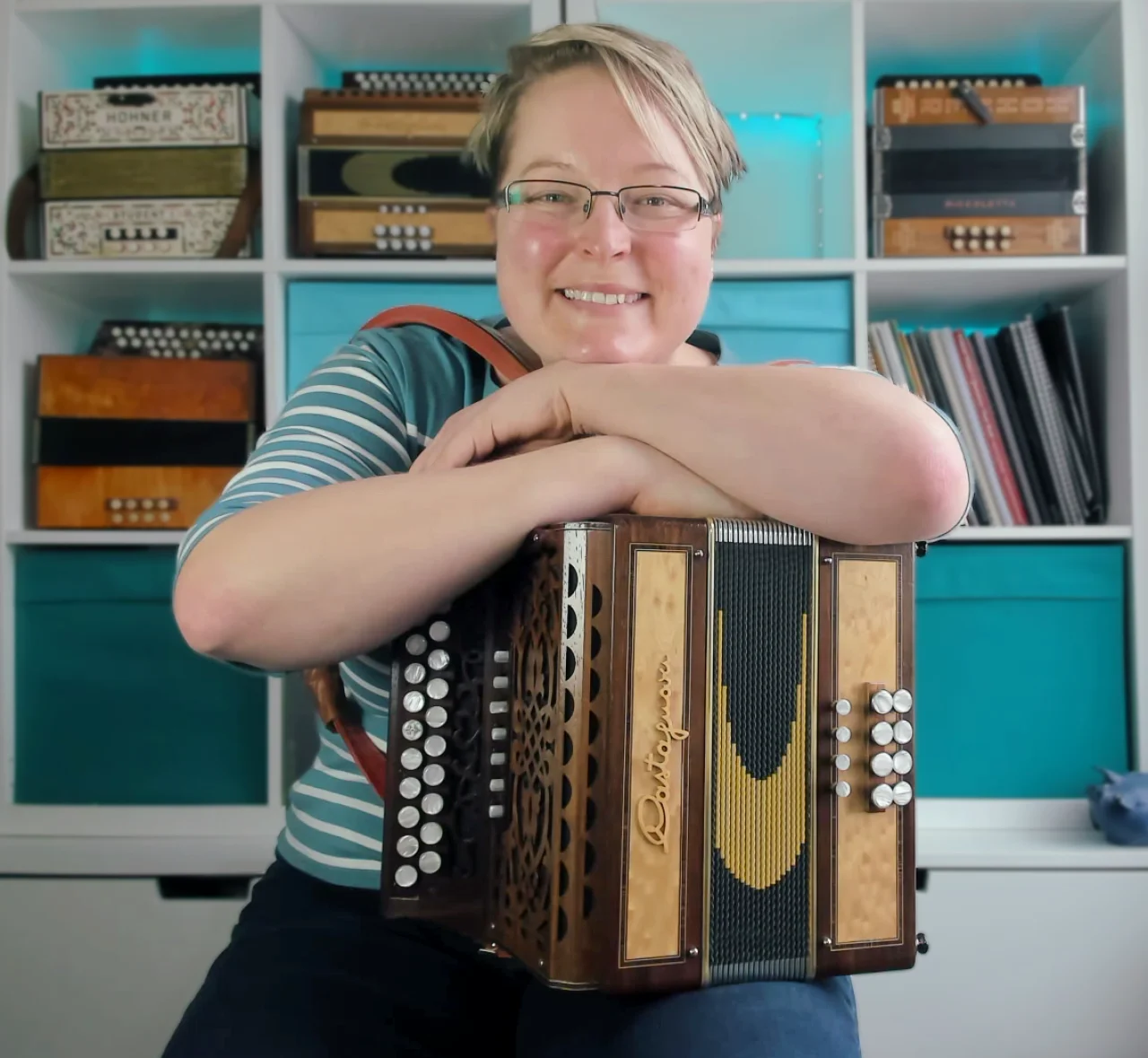 A very smiley lady with short blonde hair and half-rim glasses sits in front of an IKEA Kallax unit. In its square storage spaces, there are four melodeons, some a4 sheet music books and some blue boxes made of fabric. There is one empty space in the unit and the melodeon which is usually here is on the woman's lap in the photo. She is wearing a turquoise striped top that matched the boxes behind her, and she has her arms folded on top of the melodeon on her lap.