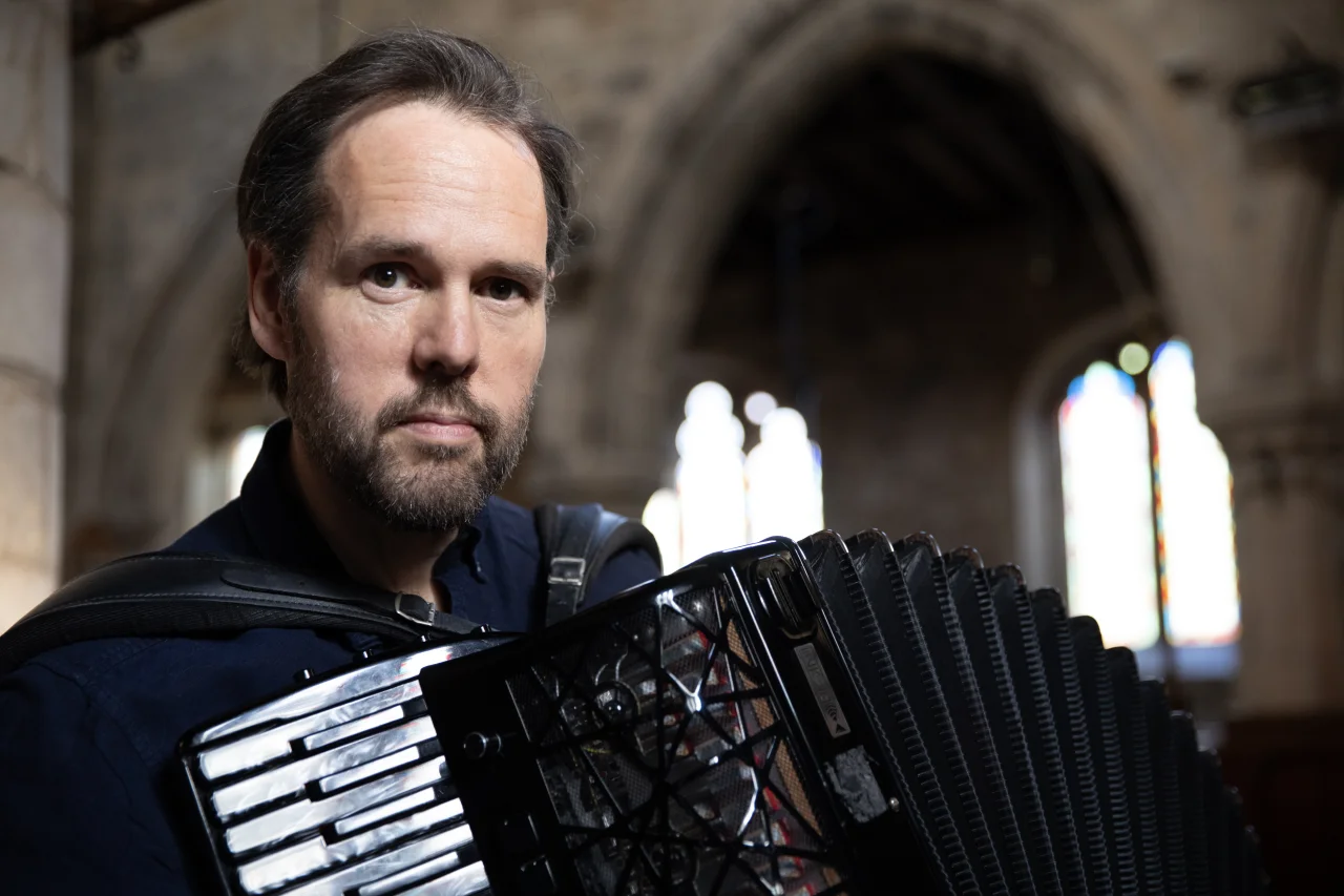 Course leader, Murray Grainger, holds his accordion and look into the camera. He has brown hair, brown eyes and a beard. He is standing in a church, the background of the photo includes arches and some blurred stained glass.