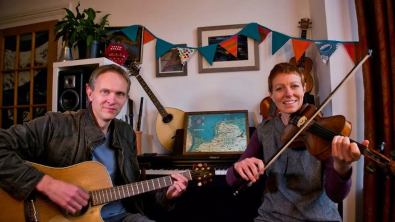 Nick Wyke & Becki Driscoll sit in a front room, in front of a piano with pictures hanging on the walls and bunting and instruments also decorating the walls. Nick wears a jacket and plays the guitar whilst looking at the camera. Becki plays the violin and also looks into the camera. Both are smiling.