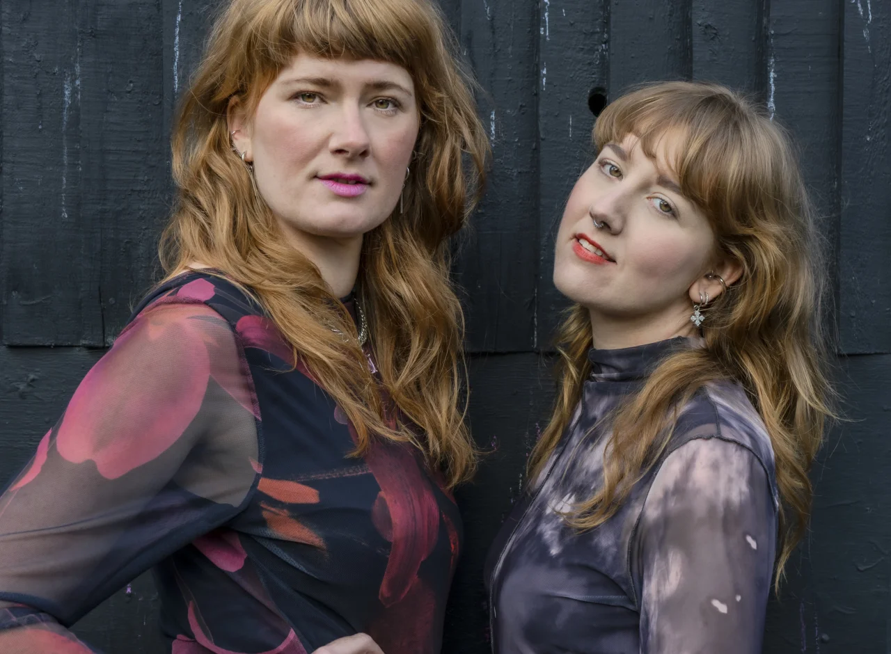 Rowan and Anna look into the camera, standing in front of a black fence. They have on purple, pink and black tops and both wear bright coloured lipstick.
