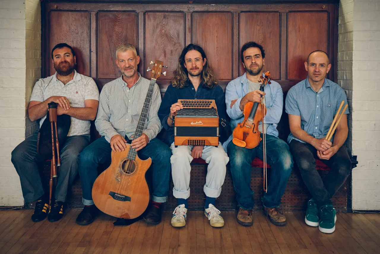 Five men sit next to each other against a wooden background. They are all holding instruments and looking at the camera.