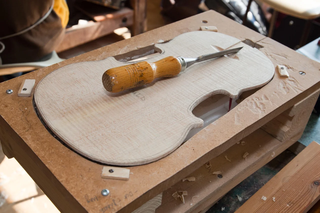 A violin mould with a violin shaped piece of wood sitting within it. A tool sits on top ready to be used.