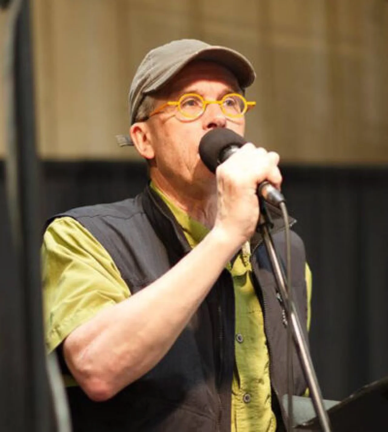 A man in a baseball cap with bright yellow round glasses holds a large microphone up to his face. He is wearing a black gilet and a lime green shirt.
