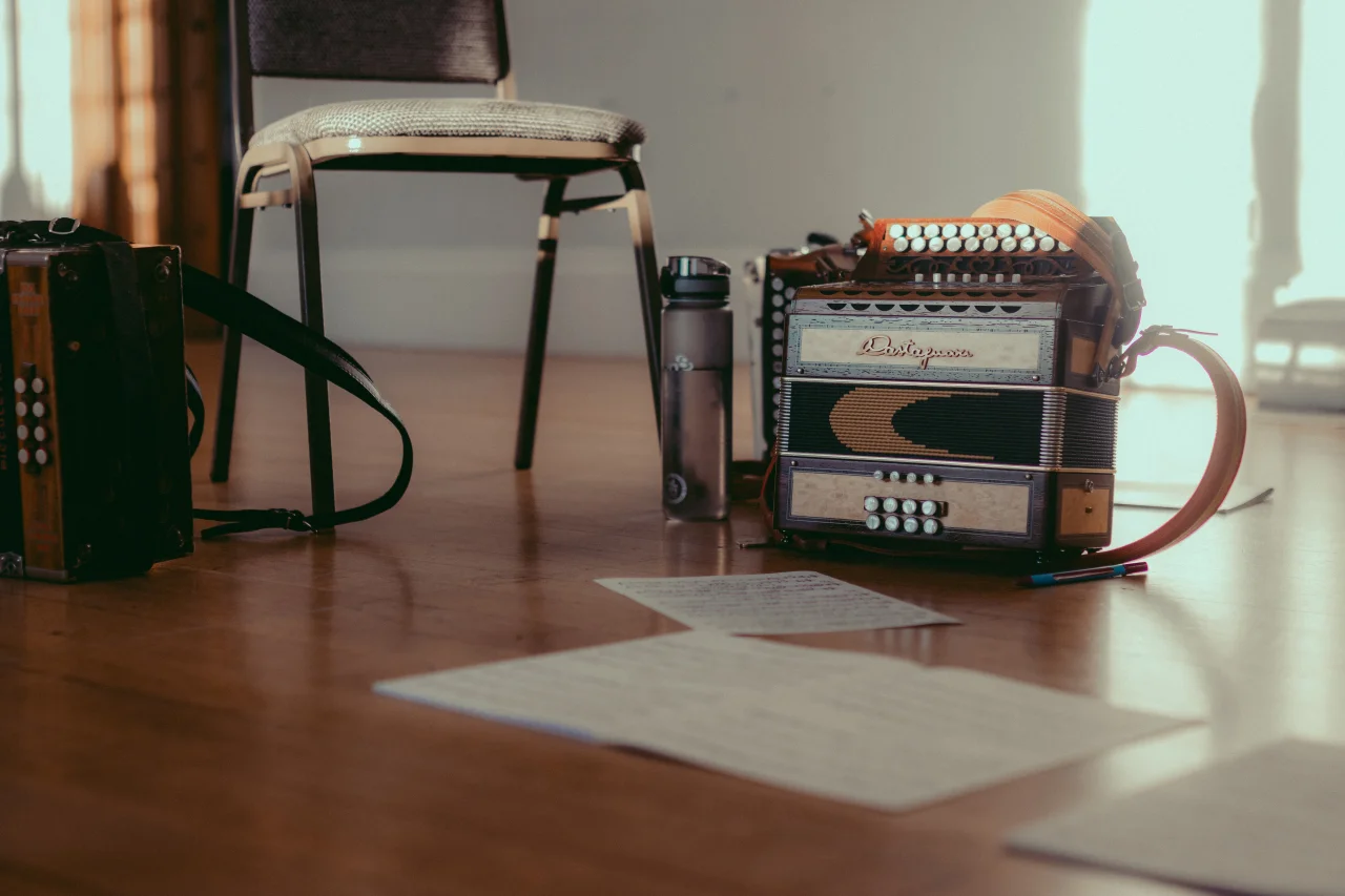 A group of objects sitting on the Long Room floor, as seen from knee height. There are two melodeons, and between them, a grey padded chair and an almost-full water bottle. There are a couple of pieces of sheet music out of focus in the foreground. There is some bright sun on the wall in the background.