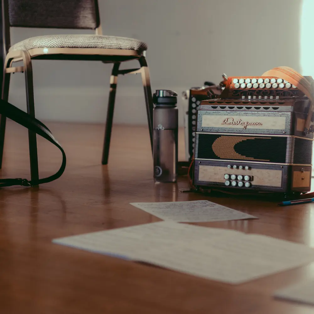 A group of objects sitting on the Long Room floor, as seen from knee height. There are two melodeons, and between them, a grey padded chair and an almost-full water bottle. There are a couple of pieces of sheet music out of focus in the foreground. There is some bright sun on the wall in the background.
