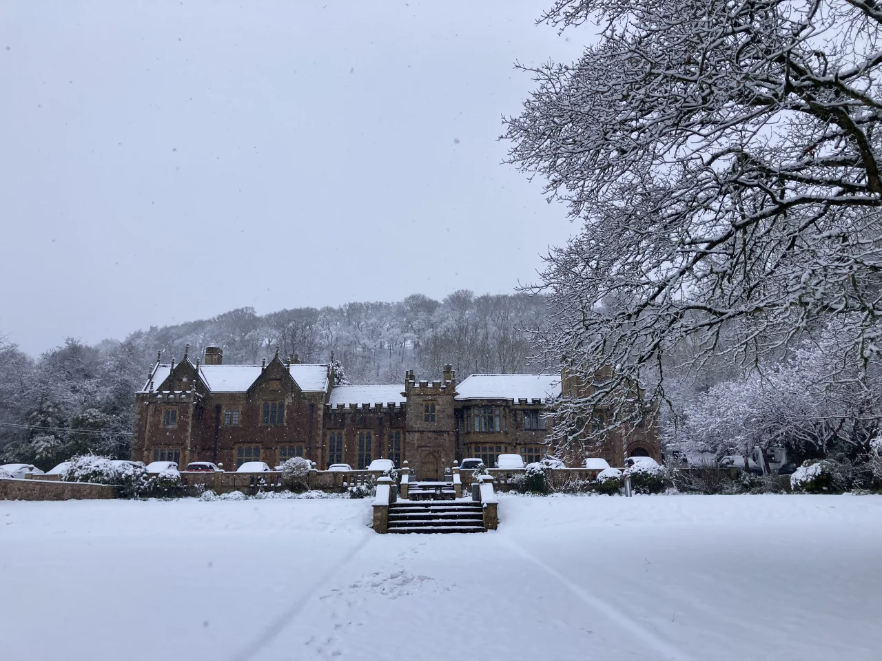 The Halsway Manor on a winters day. Snow blankets the ground and the roof of the manor. The tree branhces which frame the picture hold snow on them. The sky is a wintery grey.