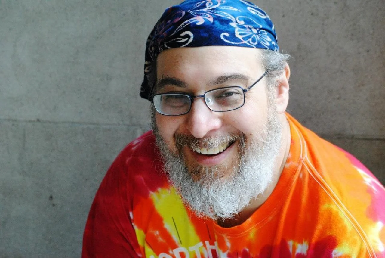 A bearded man in a bright tie-dye t shirt, blue bandana and glasses smiles broadly up at the camera.