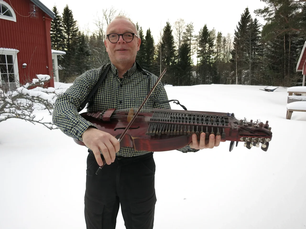 Torbjorn is standing in the snow outside a dark red scandinavian style house. There are fir trees in the background and he looks quite cold. He is holding in the middle of the frame, looking at the camera and he is wearing thick, round glasses, a dark green checked shirt and black cargo trousers. He is holding his nyckelharpa in front of him and looking right at the camera.