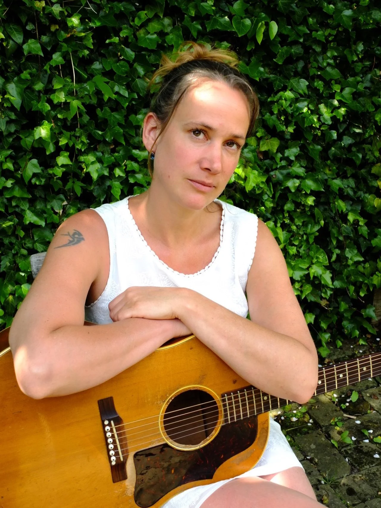 A woman in a white sleeveless dress has a guitar resting on her lap and her arms folded across the top of the instrument. She is sitting in front of an ivy covered backdrop. She has light brown hair which is held up at the back of her head, and she has a tattoo at the top of one arm.