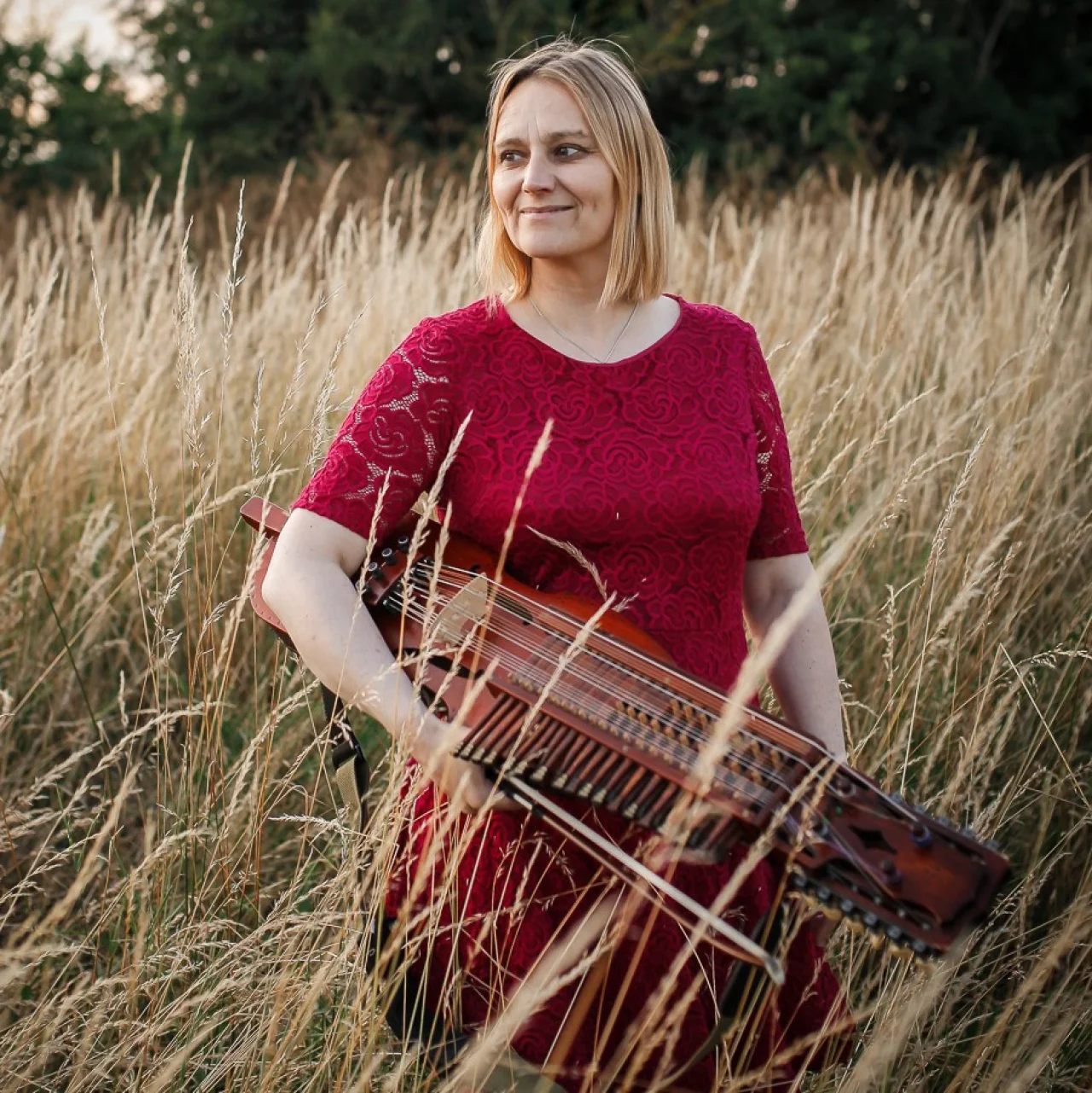 Vicki is standing in a field of very long feathery grasses and holding her nyckelharpa under her arm. She is wearing a red lace dress with elbow-length sleeves. Her hair is blond and cut in a shoulder-length bob. She is looking off to the left of the camera and smiling.